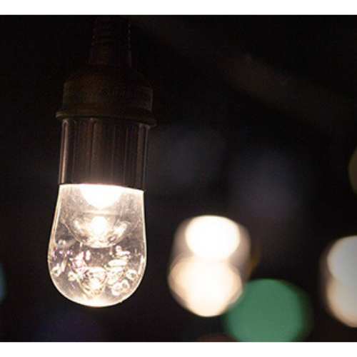A bright white light bulb hangs from a black cord in front of other blurred white outdoor lights